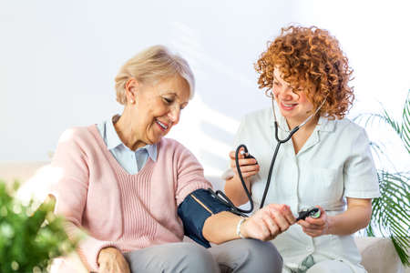 Happy senior woman having her blood pressure measured in a nursing home by her caregiver. happy nurse measuring blood pressure of a senior woman in living room Фото со стока