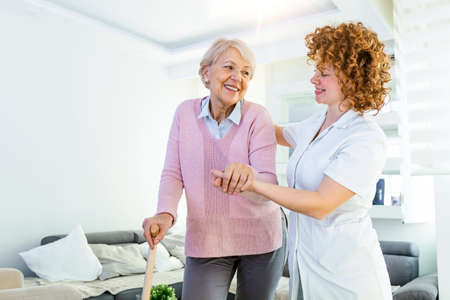 Young carer supporting senior disabled woman with walking stick. portrait of happy female caregiver and senior woman walking together at home. professional caregiver taking care of elderly woman.