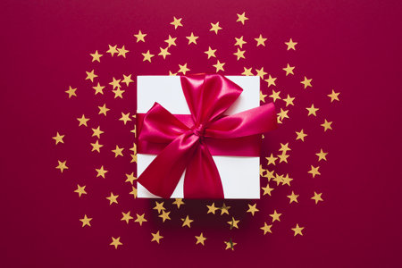 White gift box with ribbon on a red with golden confetti in the form of stars. Valentines, Christmas or New Year day backdrop. Flat lay style with minimalistic design.