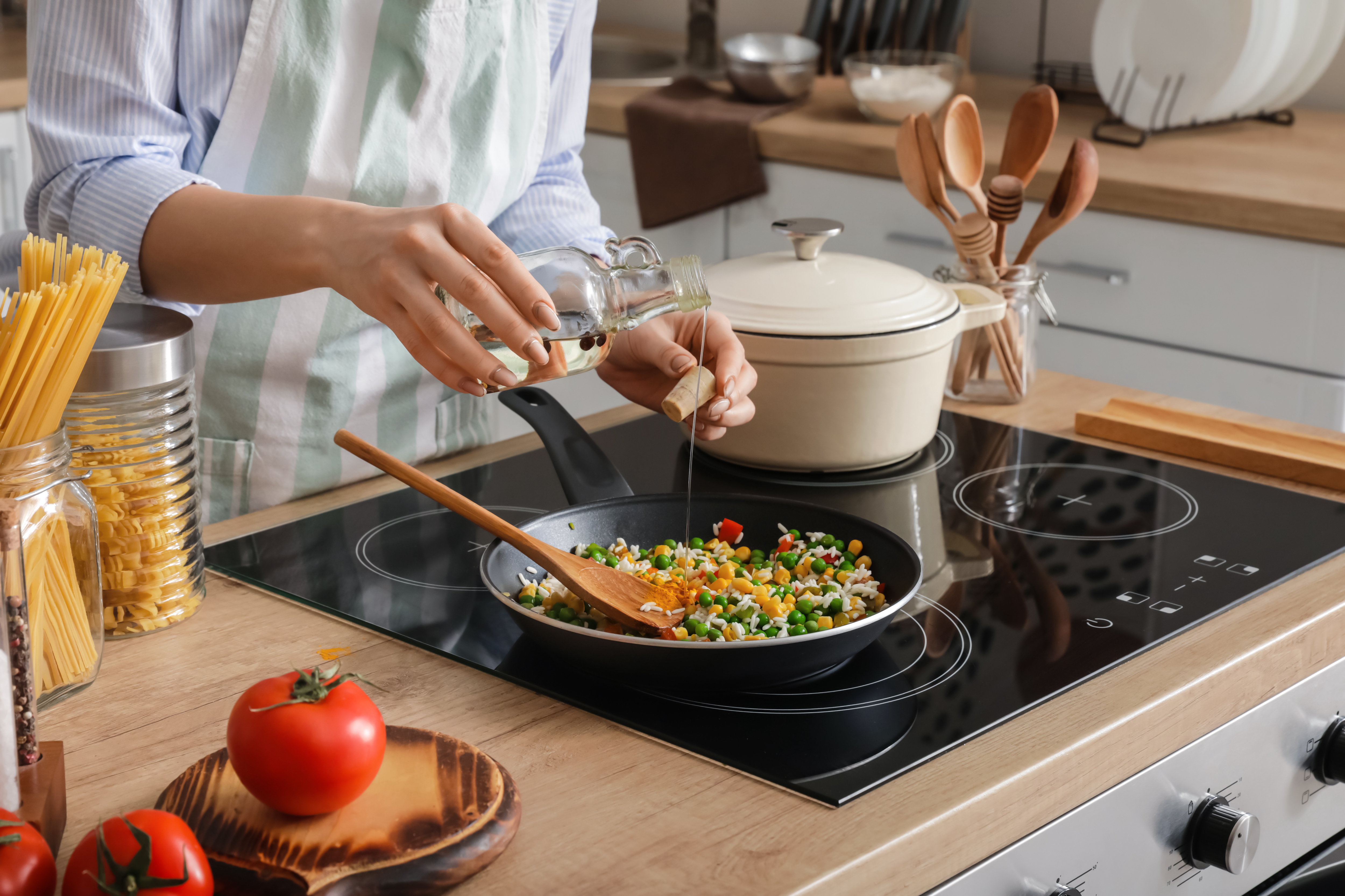 Home cook with apron pouring oil from a bottle into a vegetable stir fry cooking on a glass stove