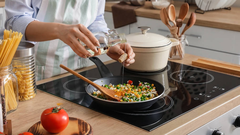 Home cook with apron pouring oil from a bottle into a vegetable stir fry cooking on a glass stove