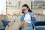 A WOMAN IN HER LIVING ROOM GESTURES IN DISMAY ON THE PHONE OVER AN OPEN SHIPPING BOX