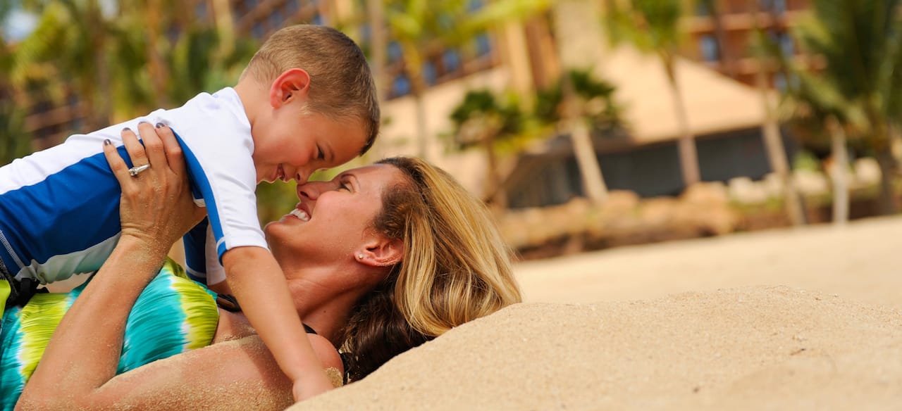 A smiling mother lies on a beach, holding her young son above her, their noses pressed together