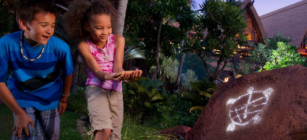 A young boy and girl discover a turtle etched into a rock hidden in trees and grass