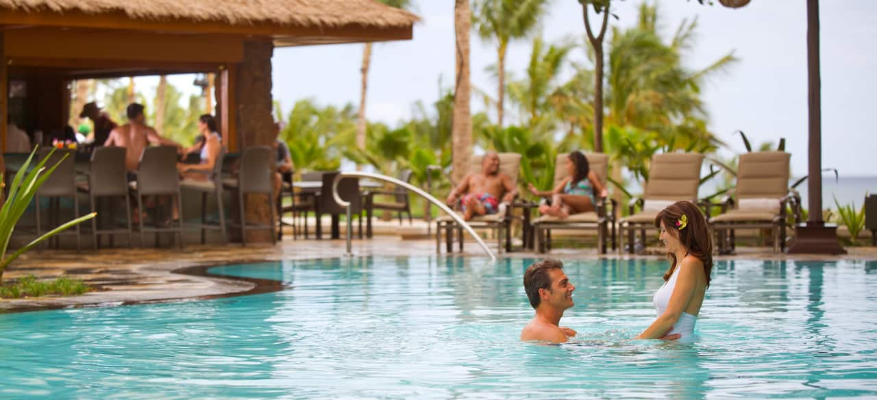 A man holds a woman by the waist in a tension-edge pool while couples lounge on chairs and drink at the bar