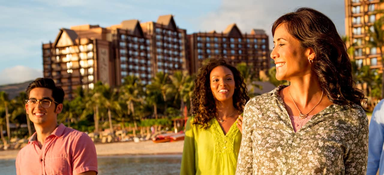 A young man and two young women walk along the beach in front of the Aulani Resort.