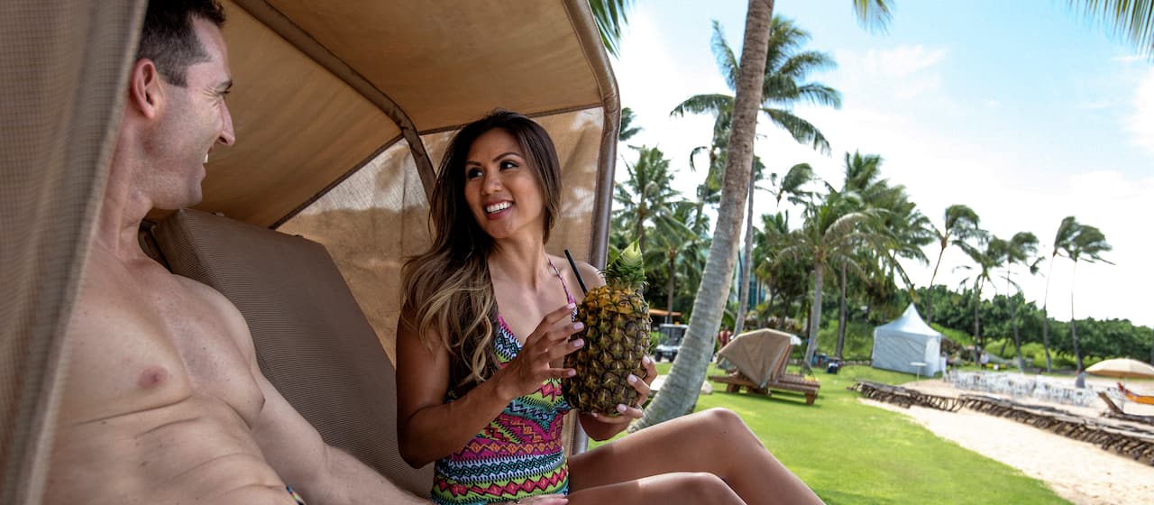 A smiling young woman holds a pineapple beverage while relaxing in a cabana with a smiling young man