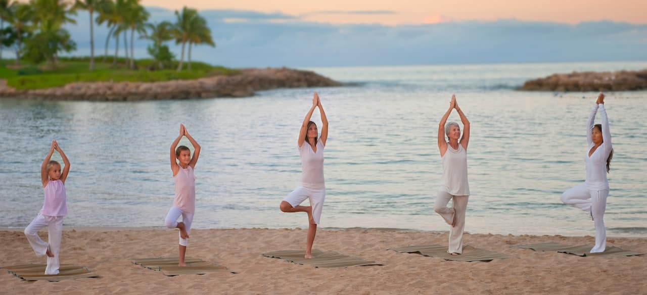 Two young girls and 3 women of various ages stand in tree pose in a yoga class on the beach