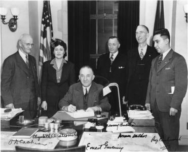 A seated Alaska Governor Ernest Gruening signs the Anti-Discrimination Act of 1945. Witnessing are (left to right): O.D. Cochran, Elizabeth Peratrovich, Edward Anderson, Norman Walker, and Roy Peratrovich.