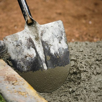 A shovel in a pool of wet cement; from the left of the frame, we see the toe of a work-boot, seemingly poised to plunge the shovel into the wet concrete.