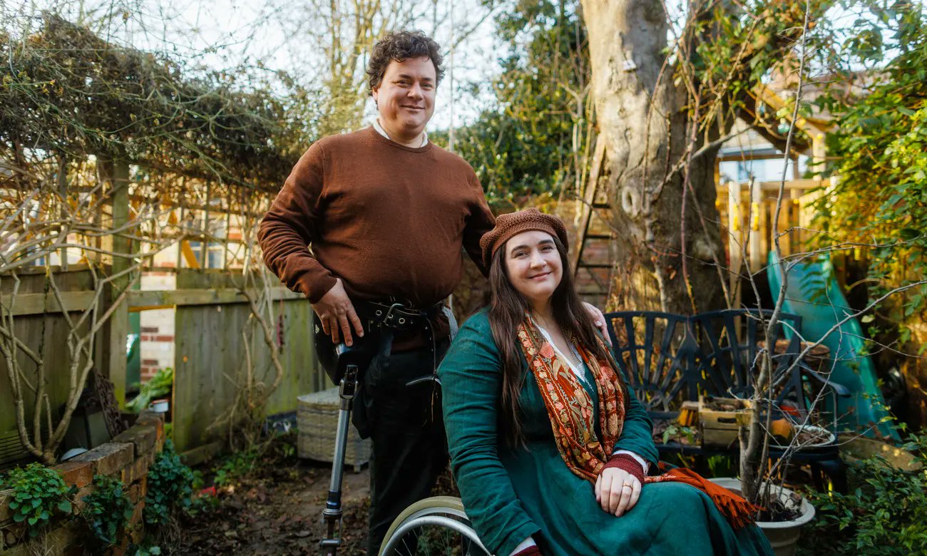 Lucy and James Catchpole in their garden. Lucy is in her wheelchair as James stands behind her.