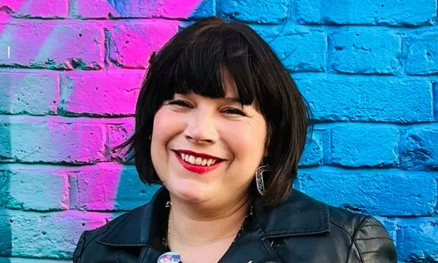 A close up of Nina Tame smiling in front of a blue and purple brick wall.