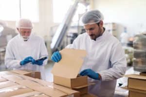 An employee in sterile uniform packing goods in boxes. In background supervisor holding tablet and counting boxes.