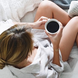 Resting girl holding a cup of coffee in bed with all grey blankets and white sheets.