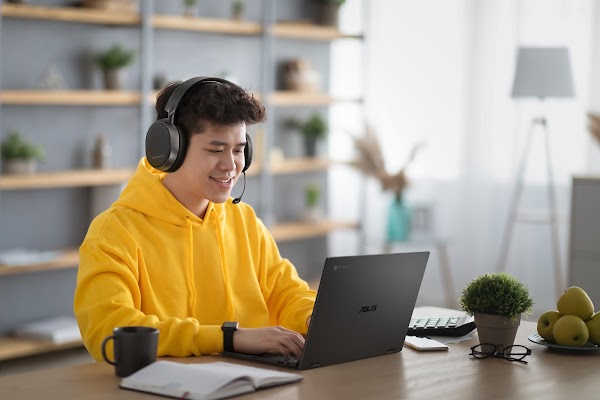 A teen gamer uses his Chromebook at a table. He wears a yellow hoodie, smart watch and gaming headset.