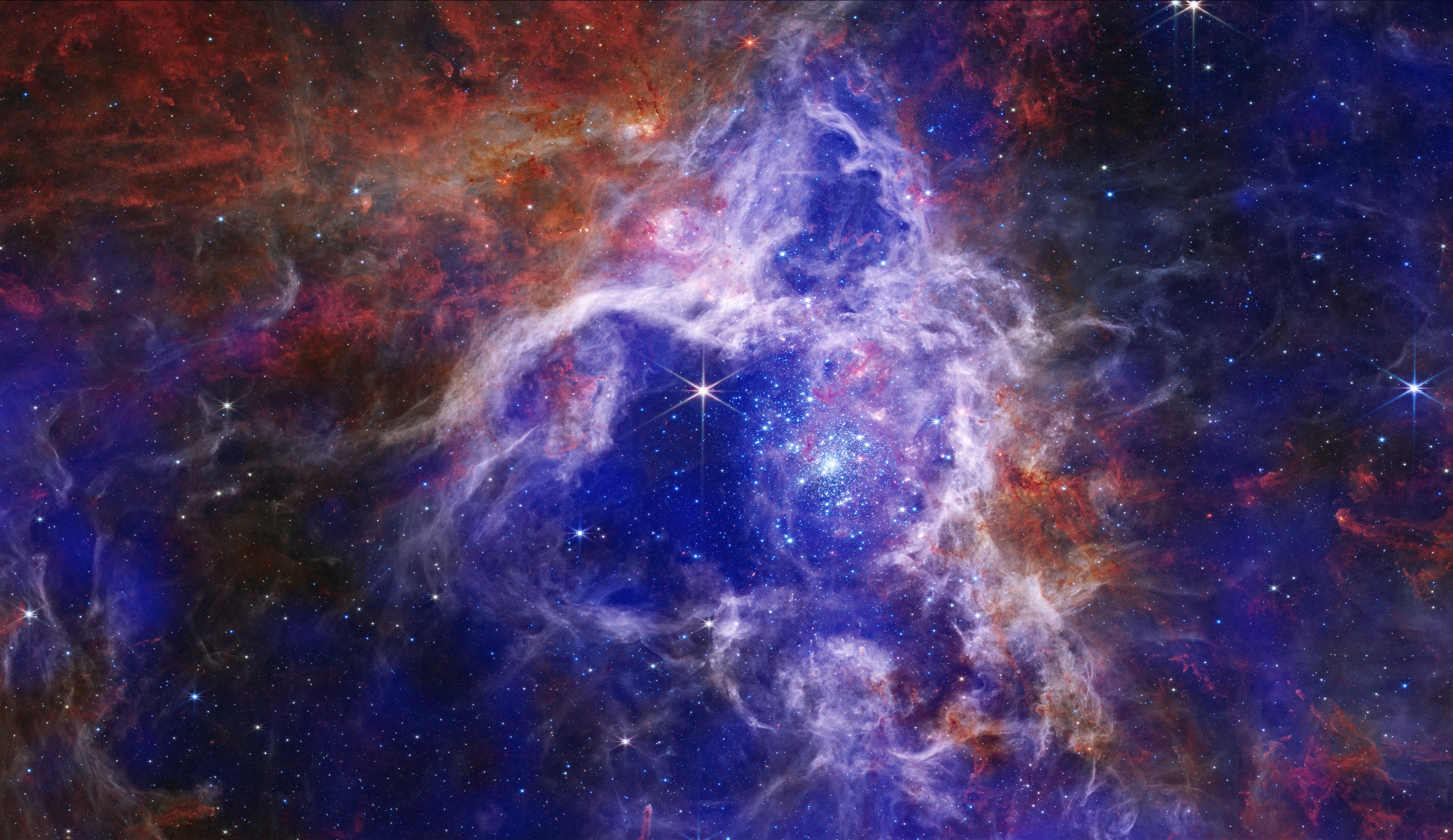 A composite image of 30 Doradus, with royal blue and purple gas clouds interact with red and orange gas clouds against a dark background. Specks of light and large gleaming stars peek through the colorful clouds.The patches of royal blue and purple gas clouds represent X-ray data collected by the Chandra Observatory. The brightest and most prominent blue cloud appears at the center of the image, roughly shaped like an upward pointing triangle. Darker X-ray clouds can be found near the right and left edges of the image. The red and orange gas clouds represent infrared data from the James Webb Space Telescope. These patches resemble clouds of roiling fire. The brightest and most prominent infrared cloud appears at our upper left, roughly shaped like a downward pointing triangle.Wispy white clouds outline the upward pointing bright blue triangle in the center of the image. Inside this frame is a brilliant gleaming star with six long, thin, diffraction spikes. Beside it is a cluster of sma