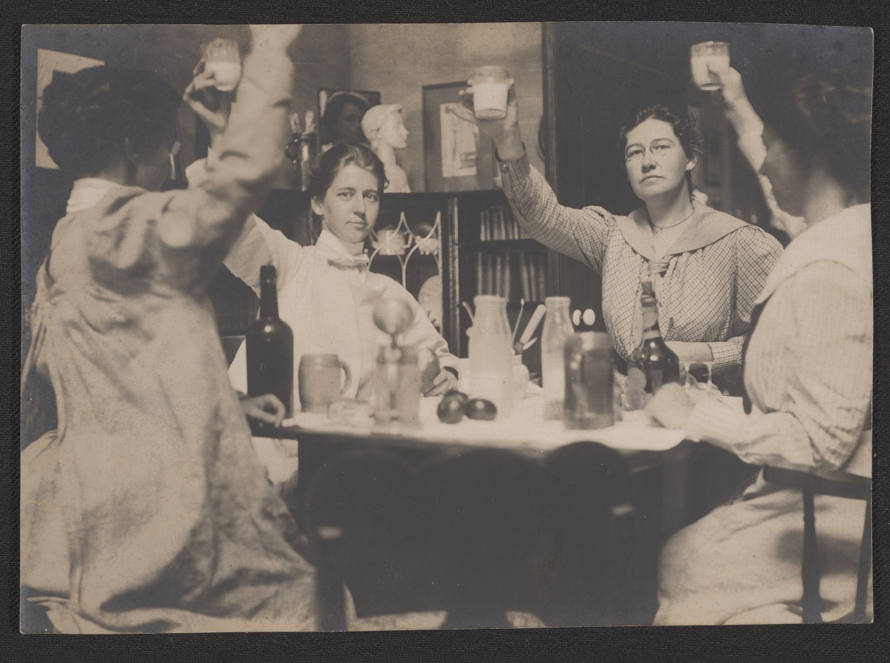 Black and white photograph of four women sitting around a table. Each woman is raising her right hand with a glass. Various bottles and pitchers sit on the table in front of them.