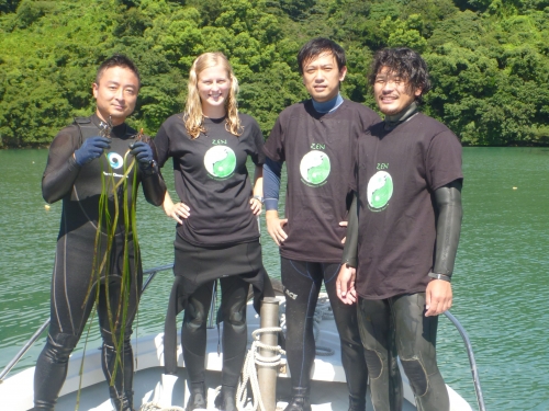 Four people in wet suits pose on edge of a boat on water