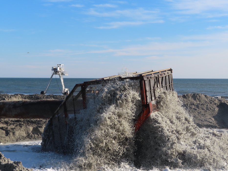 Sand and water is pumped rapidly through a pipe and screened through basket and onto a beach.