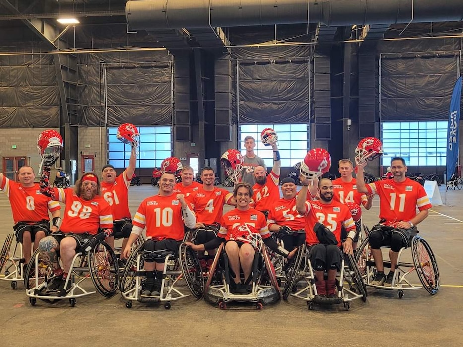 A team of men in wheelchairs pose for a photo with their helmets raised in the air in a fieldhouse.