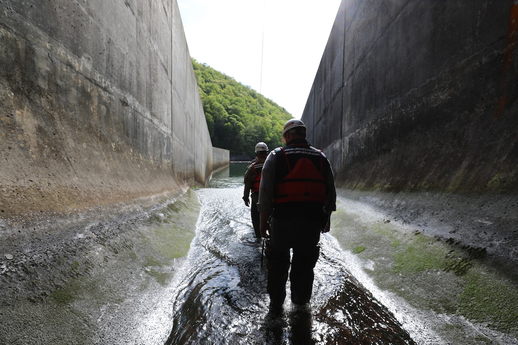 Two men walk out of a dam's outlet tunnel. The tunnel walls rise high above the men on their left and right.