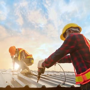 Two roofing workers in protective uniforms and wearing gloves, using electric tools and installing ironing metal sheets on top of a new roof