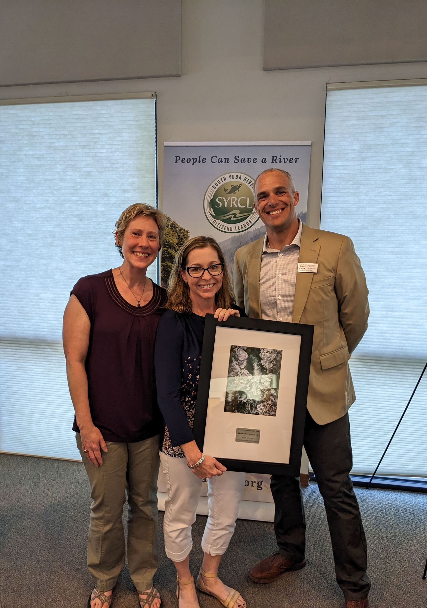 Two women and a man pose for a photo while holding a framed photo of a river. 