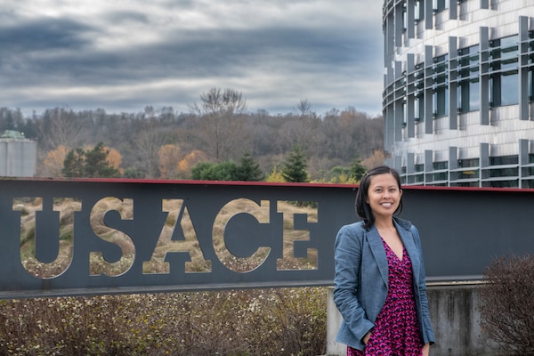 A woman in a blue jacket and maroon dress stands in front of a gray sigh with the letters USACE on it. She is standing next to a white building with window, next to a forest in fall on a cloudy day.