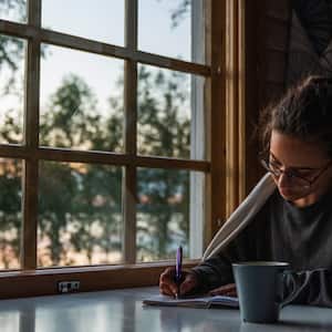woman with pen, paper, and cup of coffee sits next to window in early morning