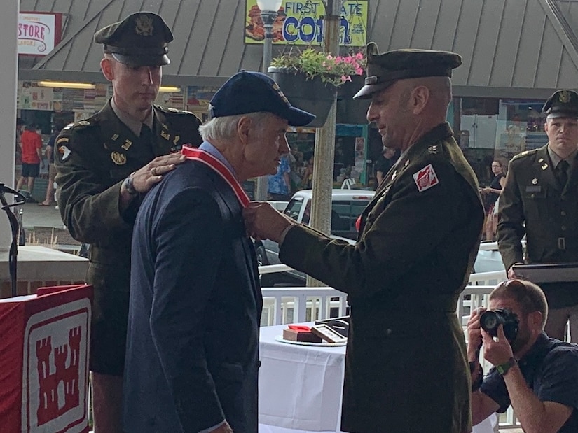 Two military members dressed in Army Uniforms present a red and gold medal to a man, Senator Tome Casper, wearing a blue hat and blue suit.