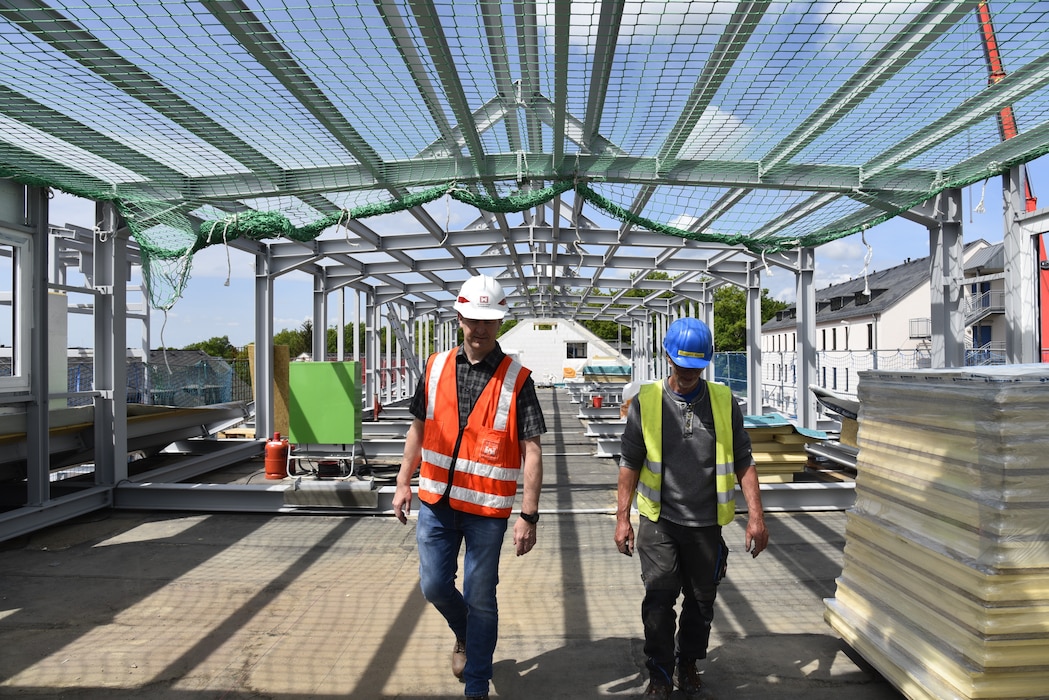 Two men in hardhats walk through a construction site. Above them a net is  strung across rows of rafters.