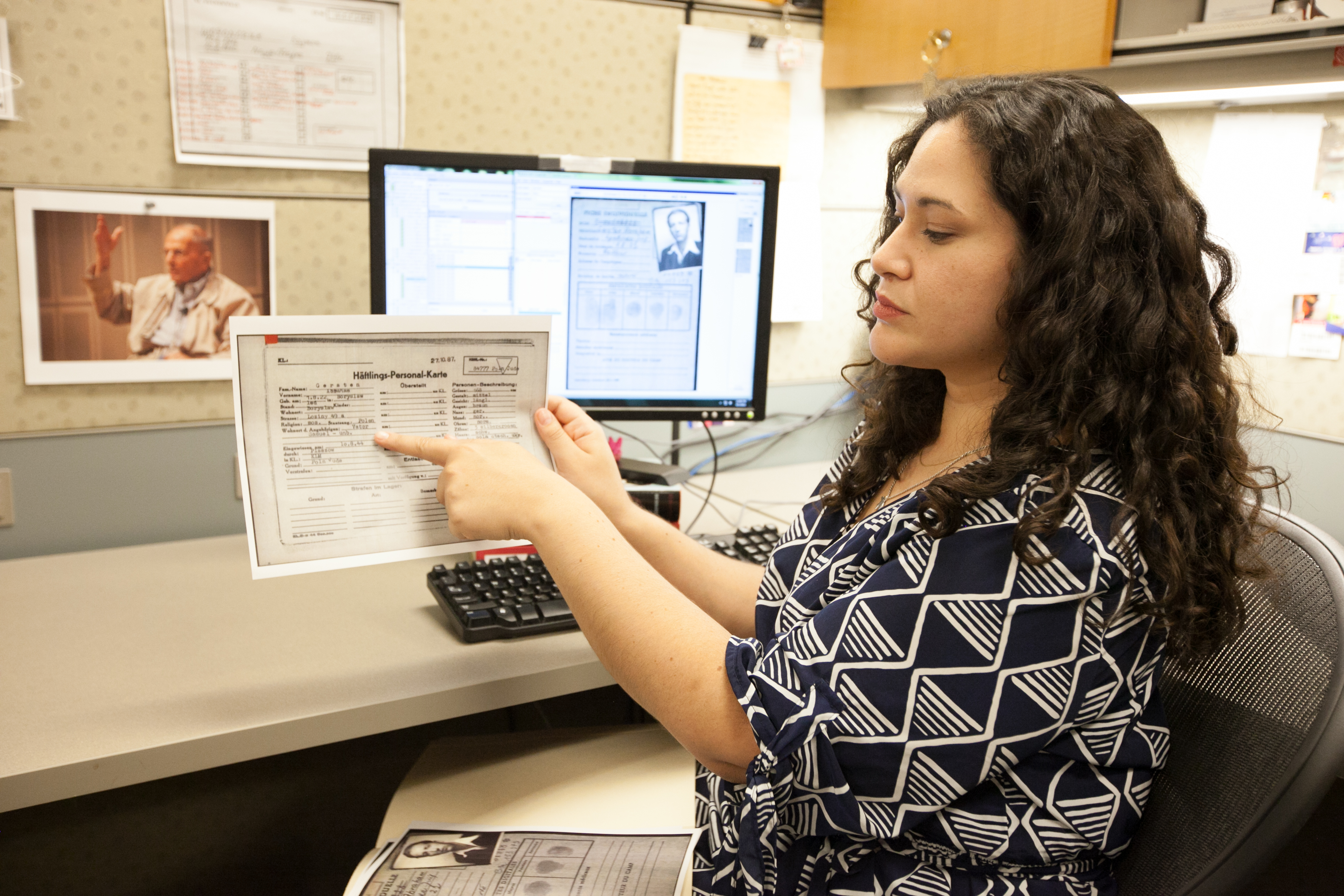 A Museum staff member holds up research while sitting in front of a computer.