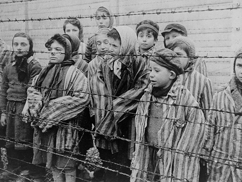 Child survivors of Auschwitz, wearing adult-size prisoner jackets, stand behind a barbed wire fence.