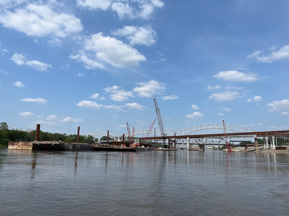 The Missouri River is shown in the foreground with construction boats and cranes in the middle, with a blue sky with clouds in the background.