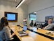 A man in a white shirt presents off of a tv screen in front of a large conference table, where three individuals can be seen sitting and listening to the presentation.