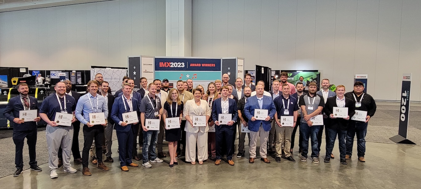 Approximately 32 people pose for a photo in a convention hall with exhibit displays in the background.