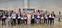 Approximately 32 people pose for a photo in a convention hall with exhibit displays in the background.