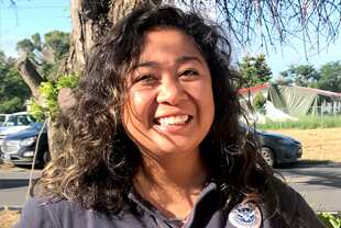 A woman smiling with trees, some cars and a damaged house behind her 