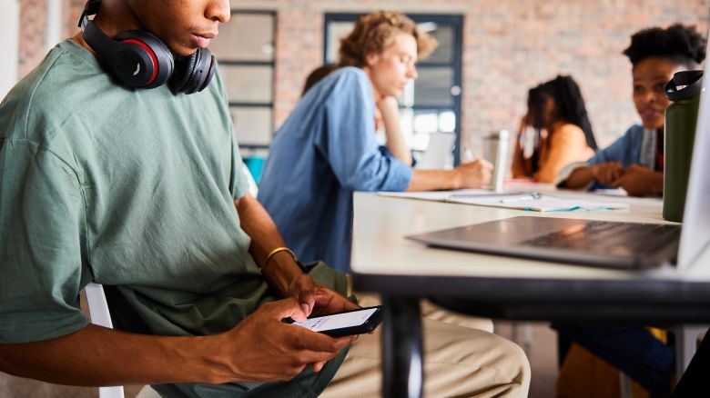 Young male student sitting at a table in a classroom during a study period and text messaging on his smart phone