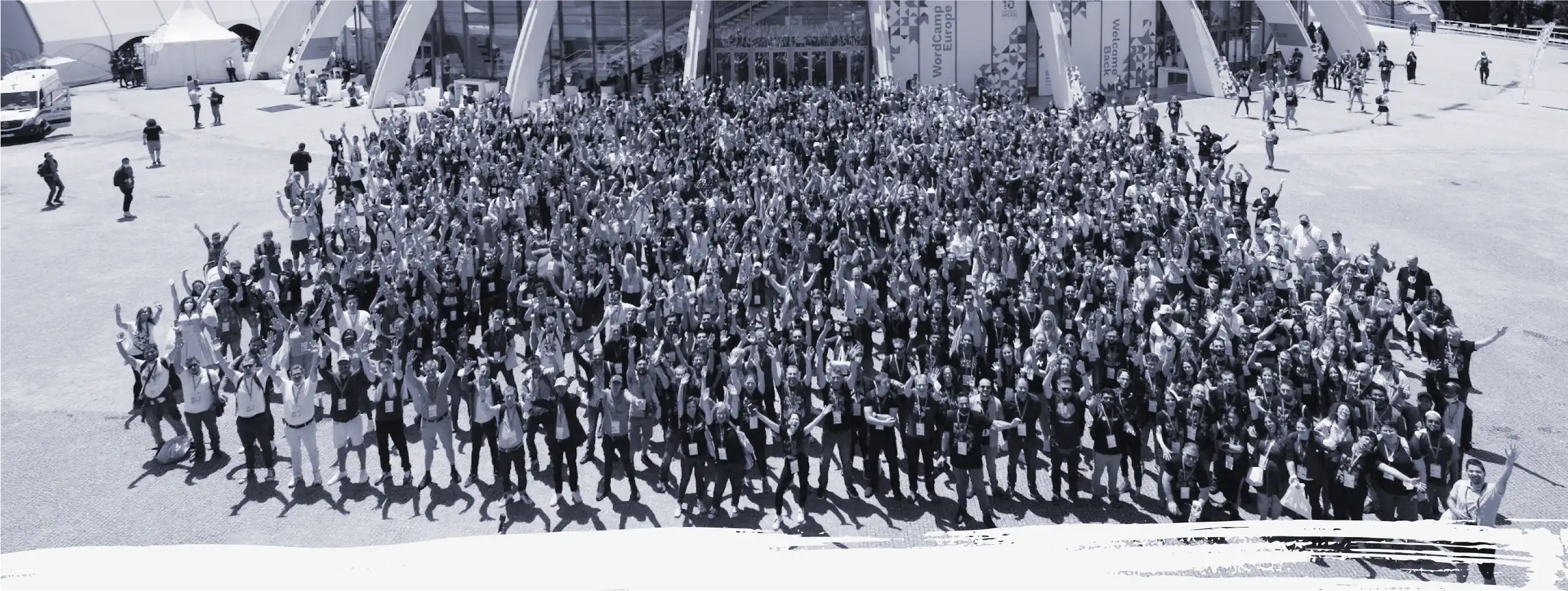 Unha vista de paxaro en branco e negro dos miles de colaboradores que asistiron a WordCamp Europa 2022. Todos eles de pé fronte ao recinto coas mans en alto en sinal de celebración.