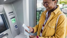 A photo of a person using a smartphone in front of an ATM while holding a debit card