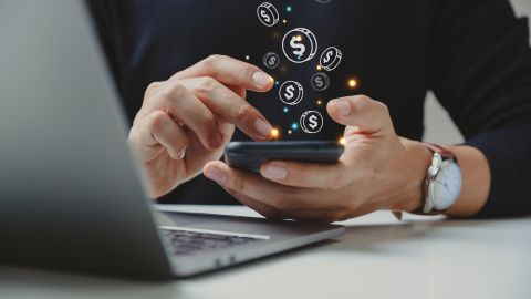 A photo of a person tapping on a smartphone in front of a MacBook with dollar signs coming out from the phone