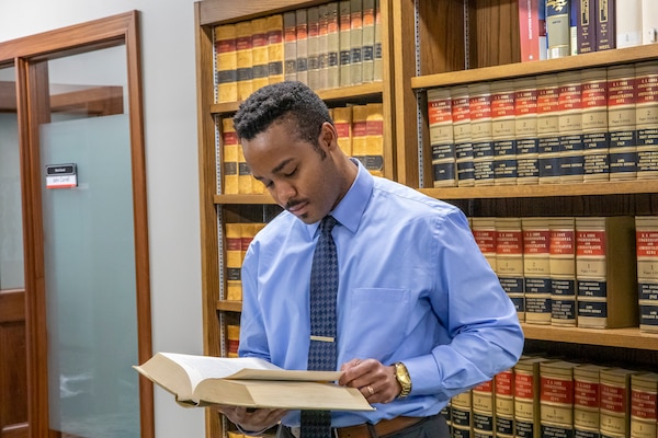 A man, Alex Webb, in a blue shirt and tie, reviews a legal book in front front of brown shelving with red, gold and black books inside of an office.