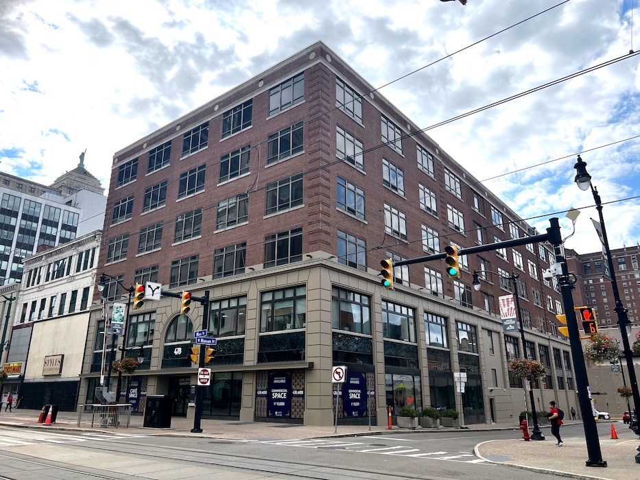 Red brick and tan building on city street corner with traffic lights and above-ground rail lines in foreground.