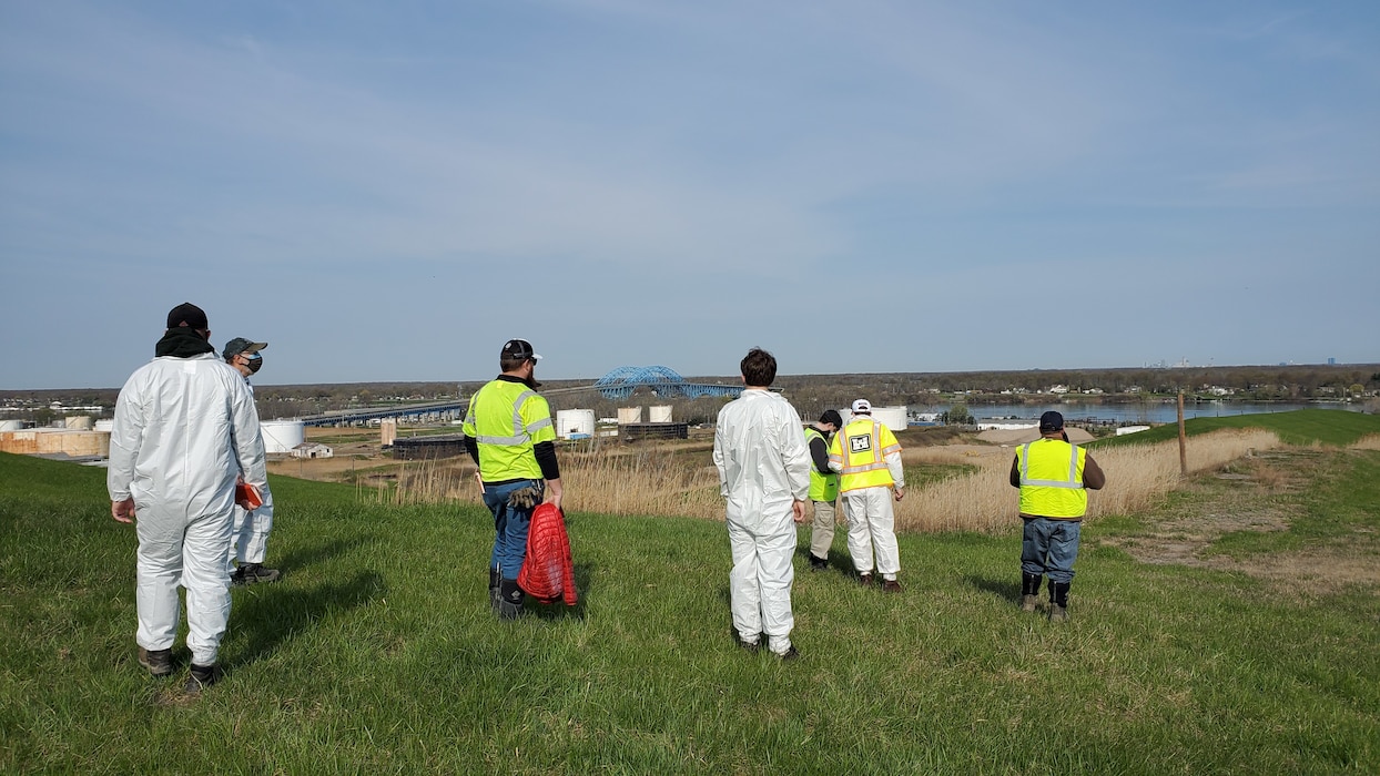 Personnel in protective clothing survey a grass-covered hill.