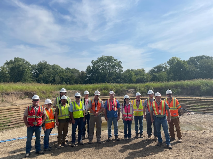 Several people in hardhats and safety vests stand in front of a creek with trees and a blue sky in the background.