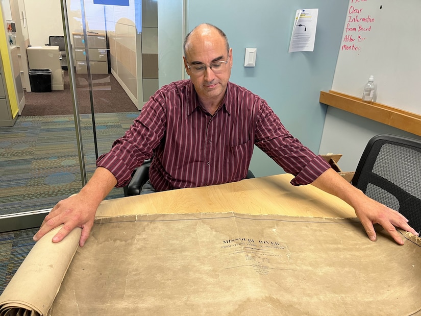 A man in a red shirt unrolls a brown sheet of paper and gazes down at it.