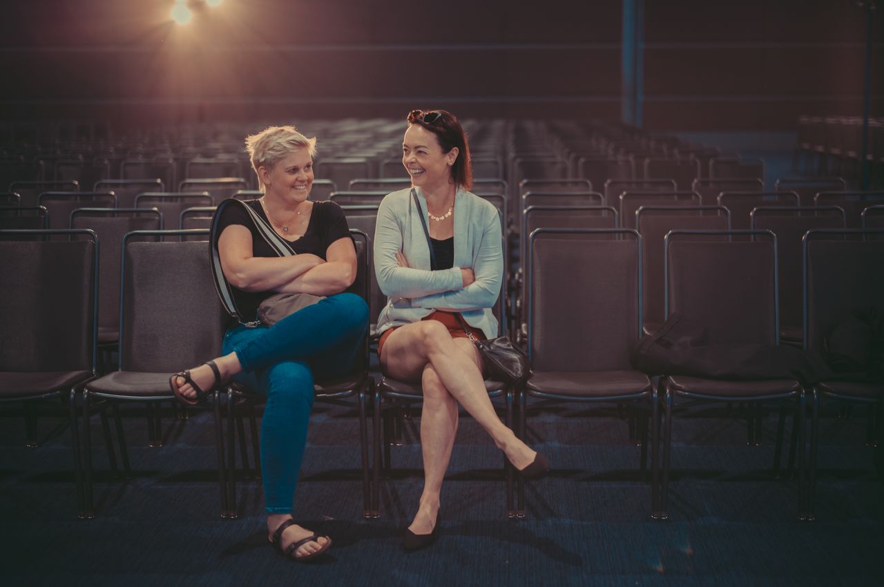 Two women sit in a large ballroom, laughing, while all the other chairs around them are empty.