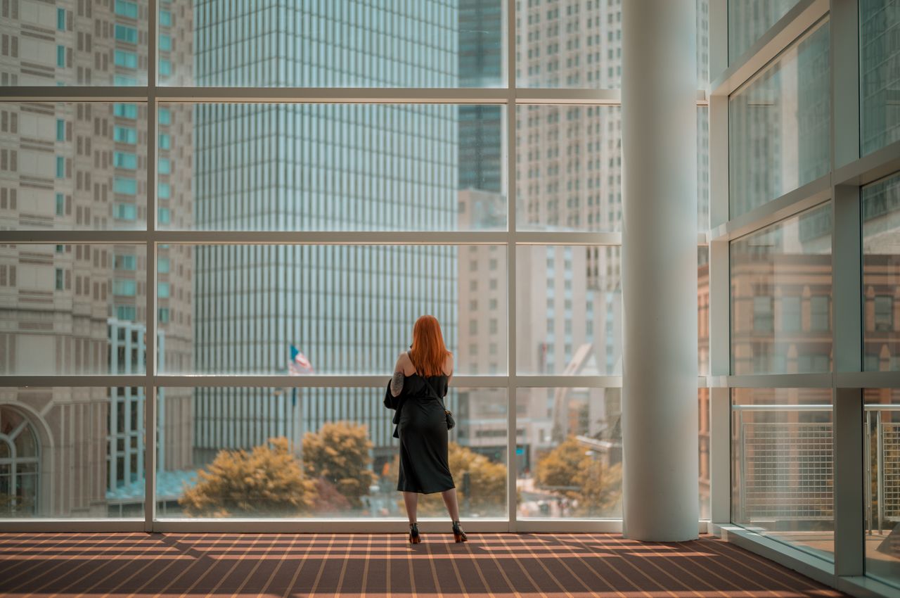 A woman stands in front of a massive window, overlooking office buildings in the city of Pittsburgh.