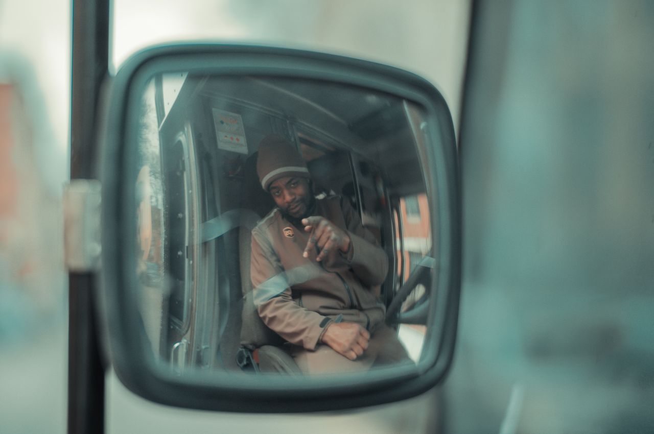 A UPS driver as seen in the side mirror of his delivery truck.